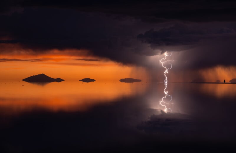 A dramatic scene of lightning striking over a vast, reflective body of water at sunset. The sky is a blend of dark storm clouds and an orange glow, with several silhouetted islands in the distance, enhancing the striking contrast of the lightning.