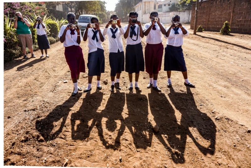 A group of seven students in uniforms stand on a dirt road, holding up cameras and taking photos. They have shadows cast on the ground in front of them. There are buildings, greenery, and another person in the background.