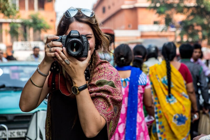 A person wearing a colorful shawl and sunglasses is holding a Canon camera and taking a picture on a busy street. Several people with vibrant clothing are visible in the background, along with buildings and trees.