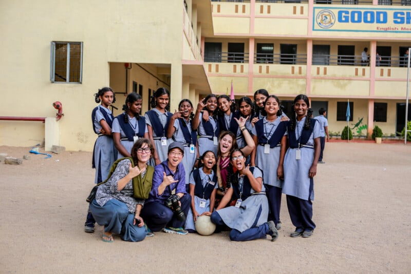 A group of students in blue and gray school uniforms pose for a cheerful photo with two adults outside a building. Some students are standing while others are squatting or sitting on the ground. A sign reading "GOOD SCHOOL" is partially visible in the background.