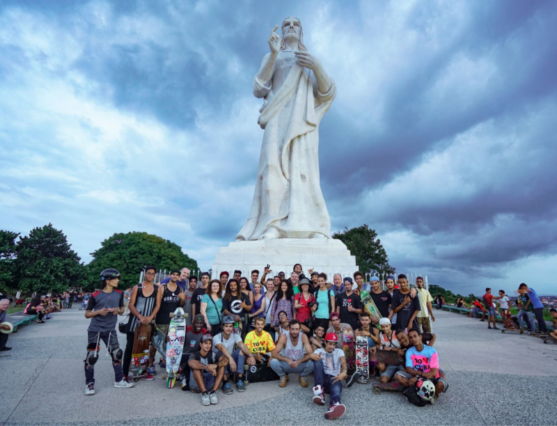 A large group of people, including skateboarders holding their skateboards, gather in front of a towering white statue of Jesus Christ. The statue is set against a dramatic, cloudy sky, and people are smiling and posing for the photo. Trees are visible in the background.