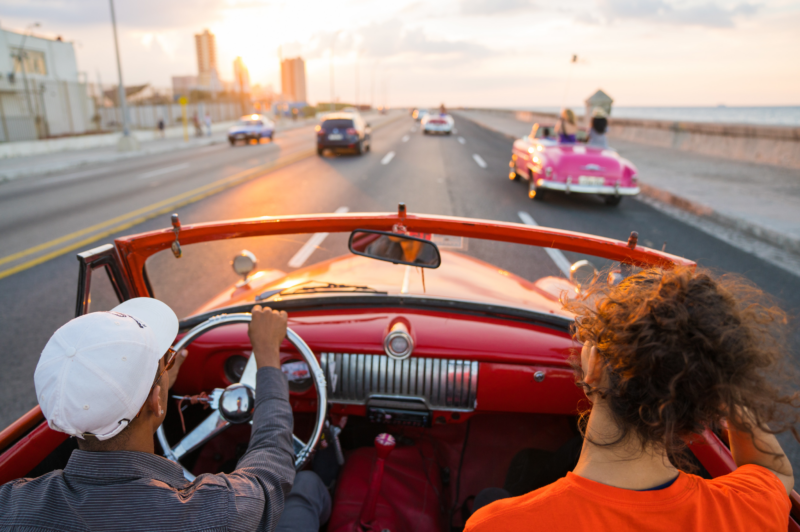 Two people ride in a red convertible along a coastal road during sunset. The driver wears a white cap and sunglasses, while the passenger has curly hair and wears an orange shirt. Other vintage cars are ahead on the road, with buildings and ocean visible in the distance.