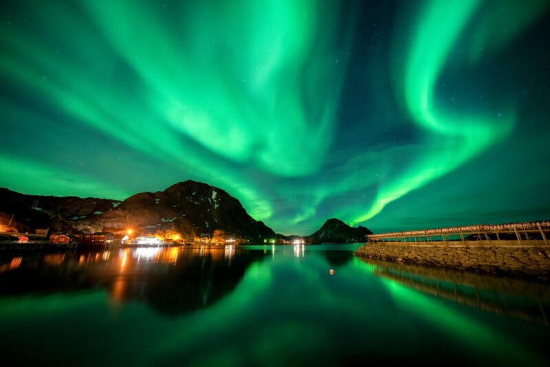 A breathtaking display of the Northern Lights illuminates the night sky with swirling green hues above a serene lake. Mountains and buildings along the shore are reflected in the calm water, adding to the enchanting scene.