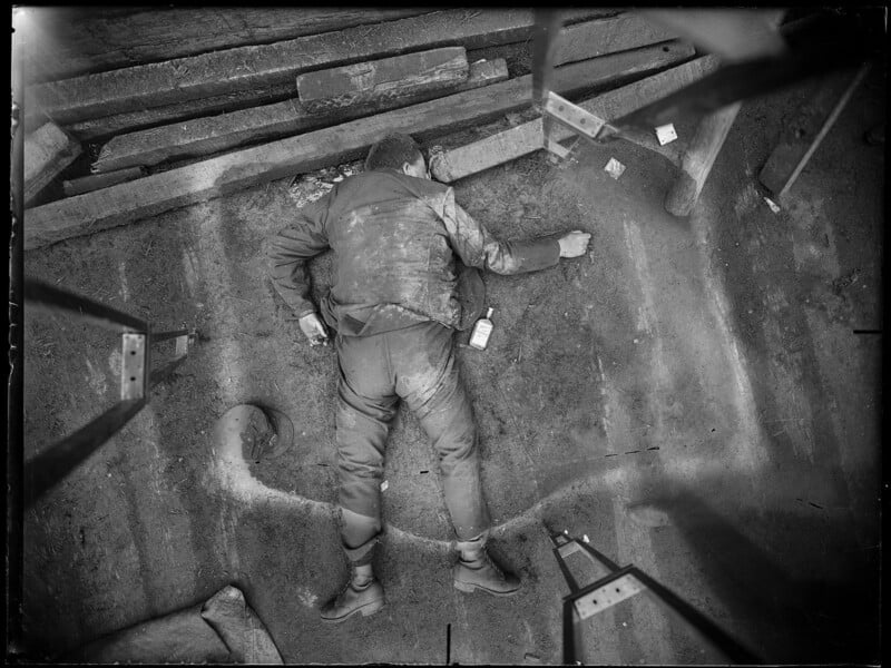 A black and white image showing an individual lying face down on the ground in a construction site. The person is wearing work clothes and appears to be surrounded by construction materials. The scene suggests a potentially serious incident or accident.