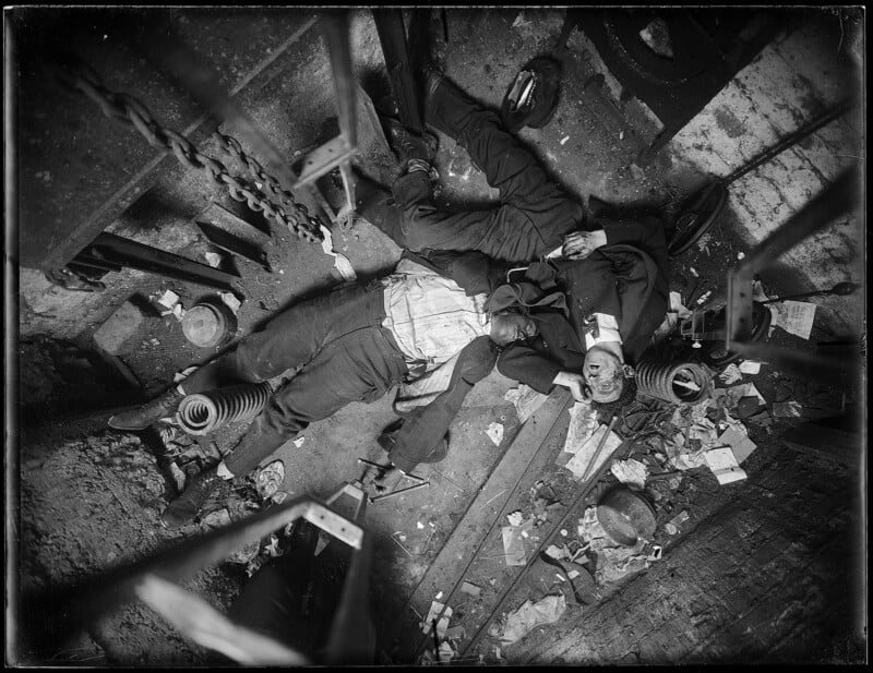 Black and white photograph of two men lying lifelessly on the floor of what appears to be an industrial setting, surrounded by scattered debris, chains, and machinery parts. The scene suggests a tragic accident, with both men wearing work attire.