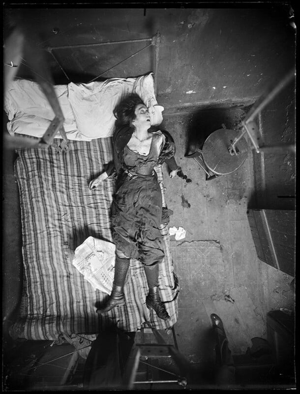 Bird's-eye view of a disheveled woman lying on a striped mattress on the floor in a cluttered room. She appears to be unconscious or sleeping, with one arm raised and her legs outstretched. Nearby, a small table holds various objects, and the floor is littered with papers.