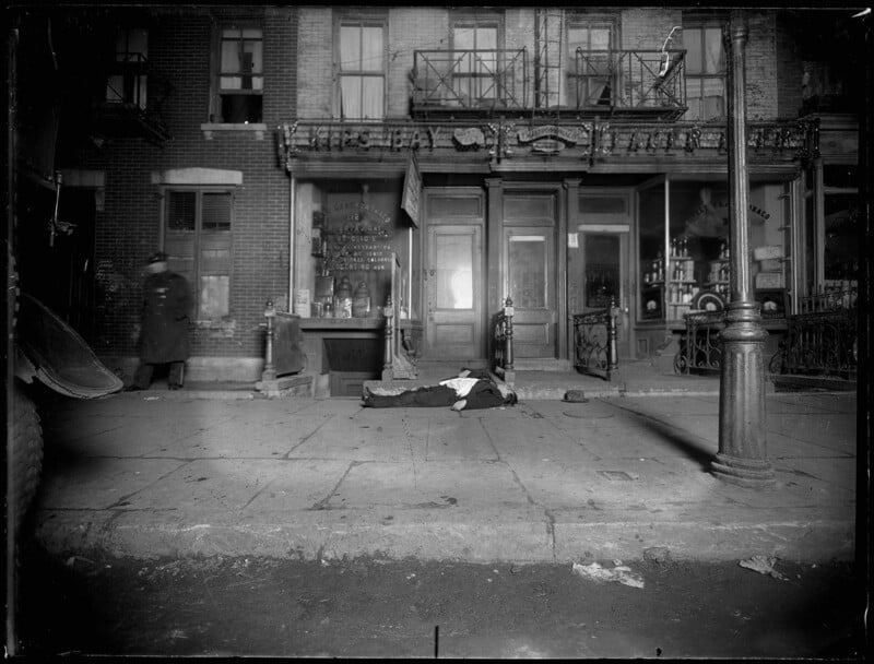 A black-and-white photograph showing a person lying motionless on the sidewalk in front of a row of storefronts. One person is walking away in the background. The image conveys a somber street scene with an air of desolation and neglect.