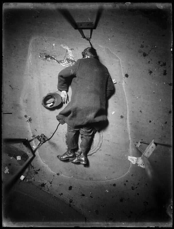 High-angle shot of a person lying on a dirty sidewalk, possibly unconscious or asleep. They are wearing a long coat and boots. A hat with a few items inside lies nearby. The scene is dimly lit, with scattered trash and debris around.