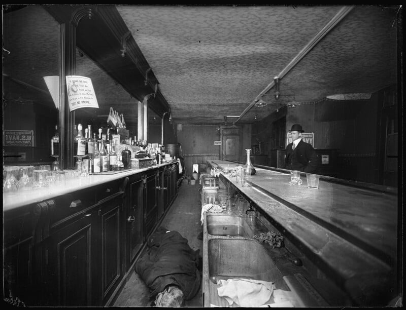 Black and white photo of an old bar interior. A man in formal attire stands behind the counter. Bottles and glasses line the bar, casting reflections. Another person lies on the floor near the counter, partially hidden, adding a dramatic element to the scene.