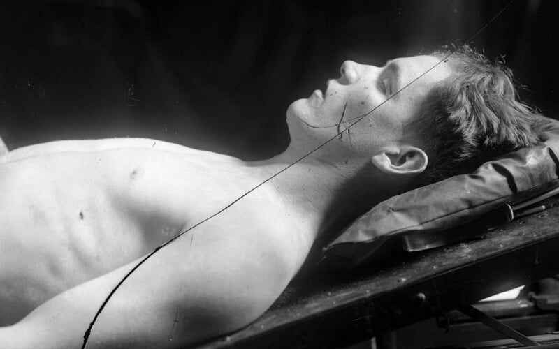 Black and white photo of a shirtless man lying on his back on a tilted platform. The man has short hair and appears to be unconscious or resting with his eyes closed. There is a visible crack in the image running diagonally.