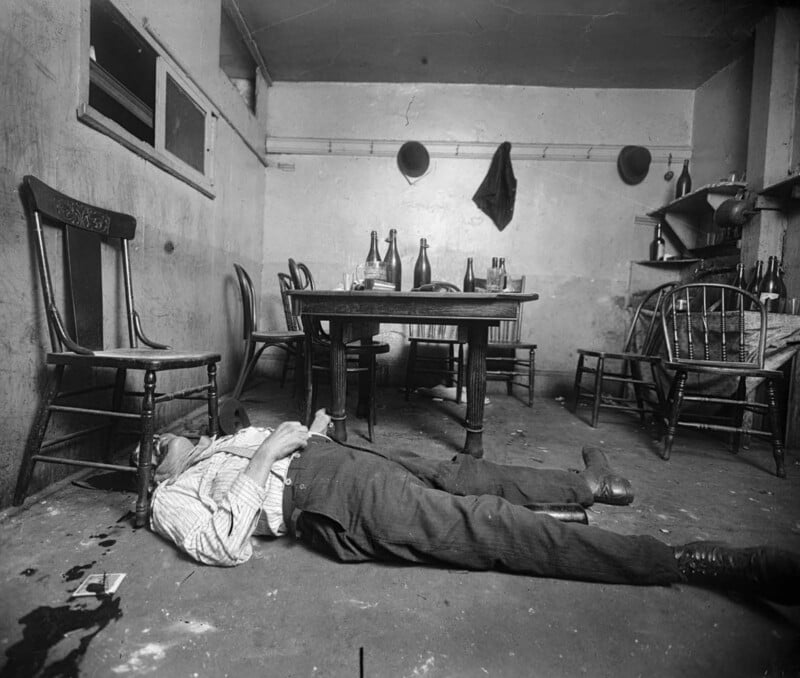 A black and white photo shows a man lying on his back on the floor of a sparsely-furnished room. The room has a table with several bottles and two chairs around it. The walls are bare and cracked, with a hat hanging on a hook and shelves with sparse items in the background.