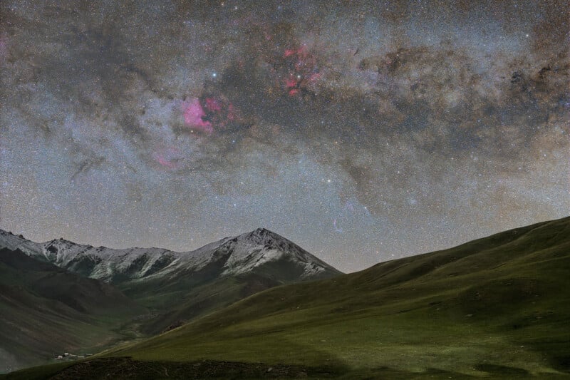 A breathtaking night sky filled with stars and vibrant pink nebulae stretches over a mountainous landscape. The foreground features green rolling hills leading to snow-capped peaks. Small buildings are nestled in the valley to the left.