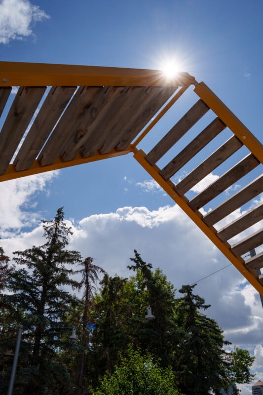 The image shows a wooden, lattice-like structure with an orange frame seen from below against a sunny, partly cloudy sky. The sun is peeking through one of the gaps in the structure's planks. In the background, there are tall trees with green foliage.