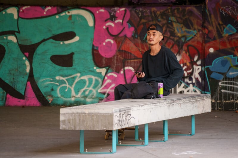 A man sits on a concrete bench in a graffiti-covered urban setting. He holds a smartphone and has a can next to him on the bench. He wears a black long-sleeve shirt, a beige cap, and dark pants, looking thoughtfully into the distance.