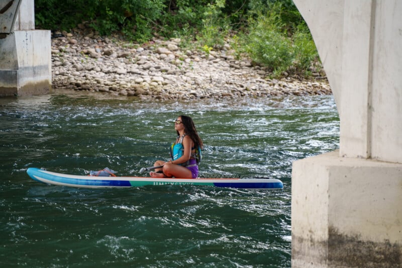 A woman sits cross-legged on a paddleboard, drifting on a river beneath a bridge. She is wearing a colorful swimsuit and sunglasses. The background features rocky riverbanks and lush green foliage.