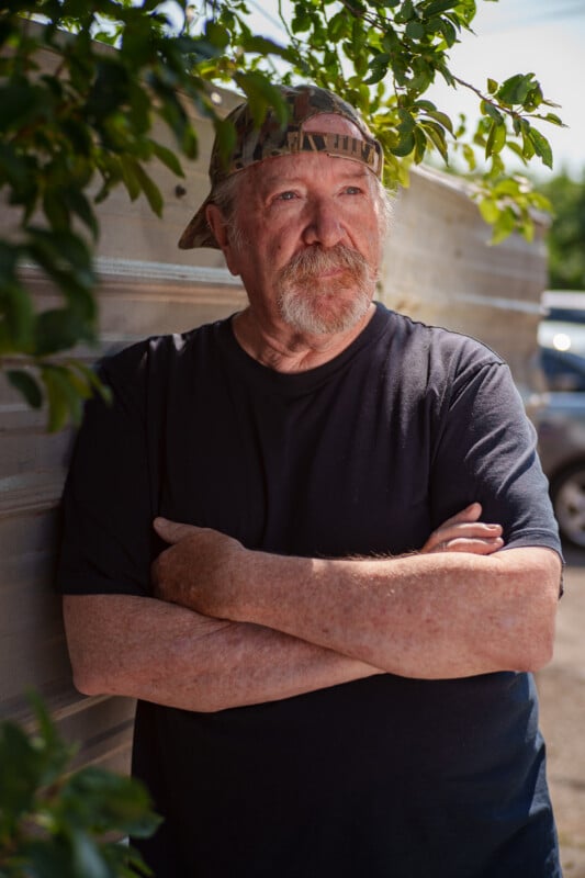 An older man with a beard and mustache, wearing a backward camouflage cap and a black t-shirt, stands outdoors with his arms crossed. He is leaning against a wooden wall surrounded by greenery and appears pensive.