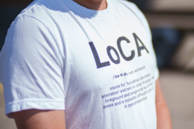 A person wearing a white T-shirt with the text "LoCA" in bold and large letters. Below "LoCA," a phonetic pronunciation and definition are provided for the acronym "longitudinal chromatic aberration," explaining its relevance to color fringes in images.