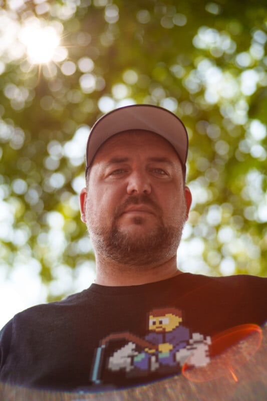 A bearded man wearing a black T-shirt with a pixel art graphic and a gray baseball cap stands outdoors. He looks down at the camera, giving a serious expression. The background is filled with bright, sunlit green foliage, creating a bokeh effect.