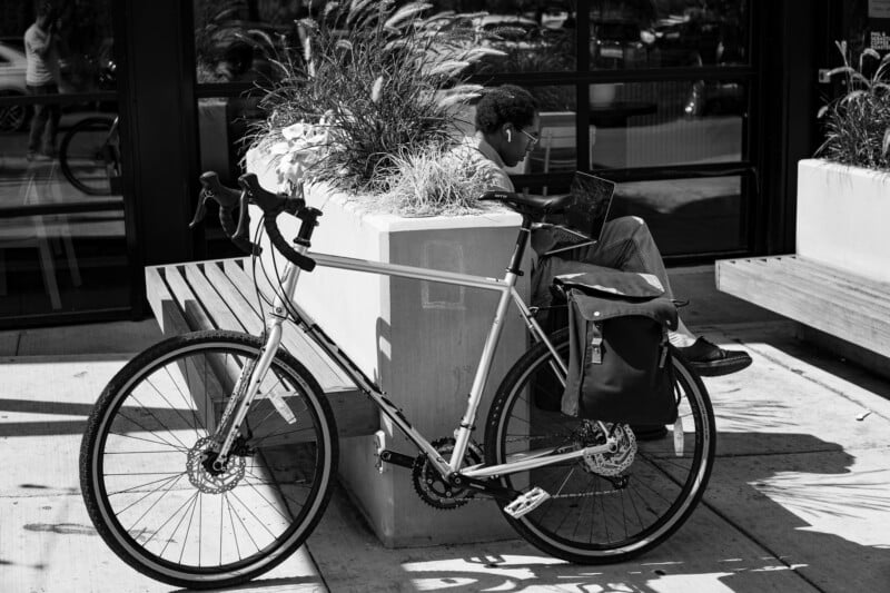 A grayscale image of a person sitting on an outdoor bench near a building, working on a laptop. A bike is parked beside the bench, with a bag attached to the back. Plants are growing in a planter box adjacent to the bench, casting shadows on the pavement.