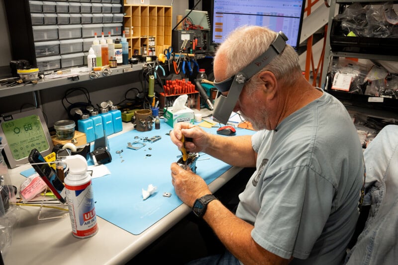 A man with gray hair, wearing magnifying glasses, works on a small mechanical piece at a cluttered desk filled with various tools, components, and containers. A computer screen displaying data is in the background. The workspace is neatly organized with shelves and bins.