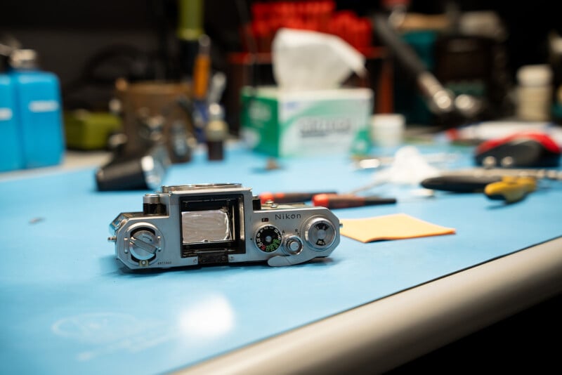 A close-up shot of a disassembled Nikon camera placed on a blue workbench. Various tools and camera parts are scattered in the background, suggesting a repair or maintenance process. The setting appears to be a workshop with a focused light illuminating the work area.