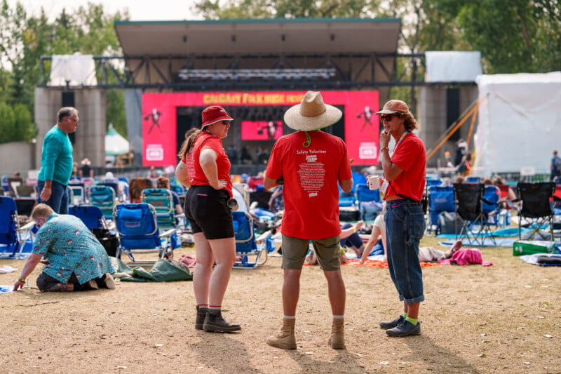 Three people, wearing matching red shirts, converse in front of an outdoor stage. The stage features a large screen displaying “Calgary Folk Music Festival.” The venue has scattered audience members sitting on the ground and lawn chairs, enjoying the event.