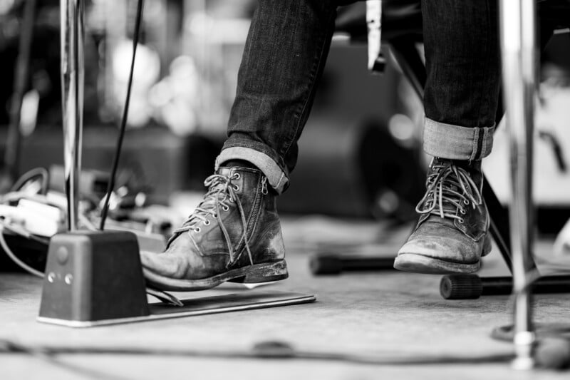 Black and white photo of a person seated, showing only their legs and worn leather boots. The boots are resting on a pedal, suggesting they are playing a musical instrument. Various cables and equipment are visible on the floor.