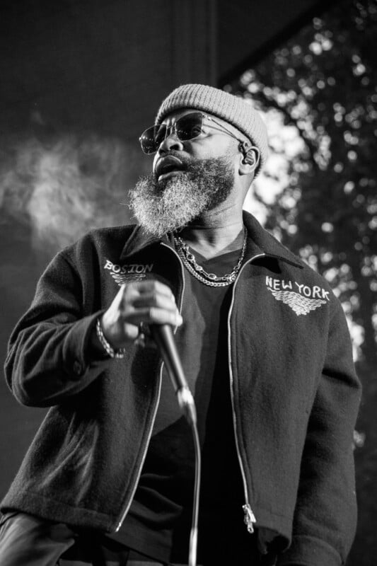 A black and white photo showing a bearded man wearing sunglasses, a beanie, and a jacket with "Boston" and "New York" stitched on it. He is holding a microphone and appears to be performing on stage, with blurred lights and trees in the background.