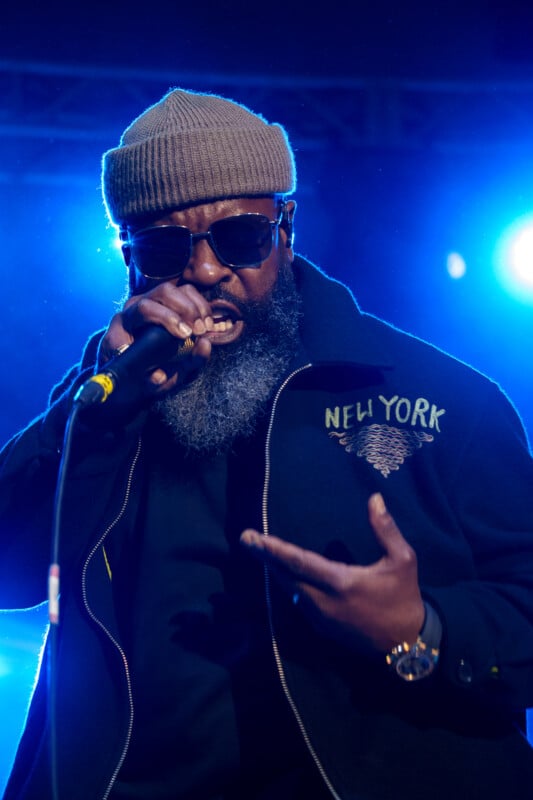 A man with a full gray beard is wearing sunglasses, a brown knit beanie, and a dark jacket with "New York" lettering. He is passionately performing into a microphone with vivid blue stage lights in the background.