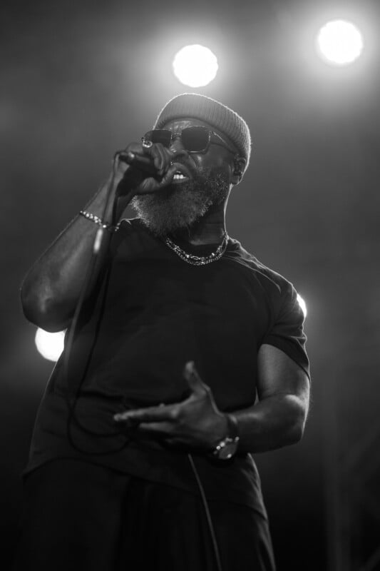 A man with a beard, wearing sunglasses and a beanie, performs passionately on stage, holding a microphone with one hand. The image is in black and white, with bright stage lights shining in the background, adding to the dramatic effect.