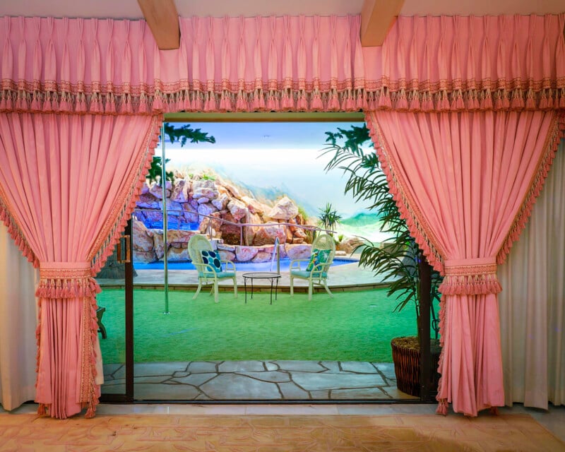 A lavish room with ornate pink curtains opens to a patio area. The outdoor scene depicts a rocky landscape mural. In the patio space, there is green flooring, two white wrought iron chairs, a small table, and a large potted plant.