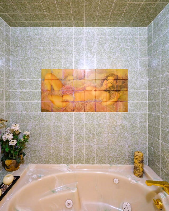 A bathroom with green tiled walls features a mural depicting a reclining woman on the back wall. The bathtub has off-white surfaces with gold fixtures. There are potted white flowers and a candle on the edge of the tub.