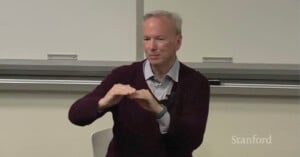 A person with a light complexion, wearing a dark sweater, sits and gestures with both hands while speaking. The background features a whiteboard and the word "Stanford" is visible in the bottom right corner.