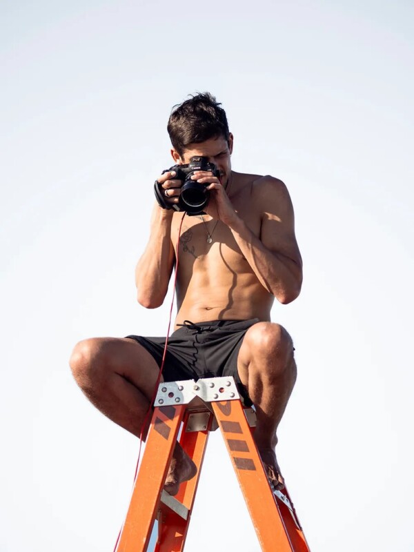 A shirtless man wearing black shorts sits on top of an orange ladder against a clear sky. He holds a camera up to his face, focusing intently as he takes a photo. His expression is concentrated and the overall scene suggests he is involved in a photography shoot.