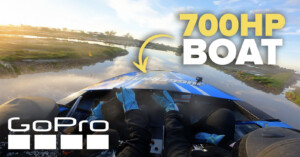 A thrilling scene captured from a GoPro shows a high-speed boat ride on a river. Two individuals are visible from behind, steering the boat. Text overlays indicate "GoPro" and "700HP BOAT." The background features a scenic sunlit landscape and reflective water.