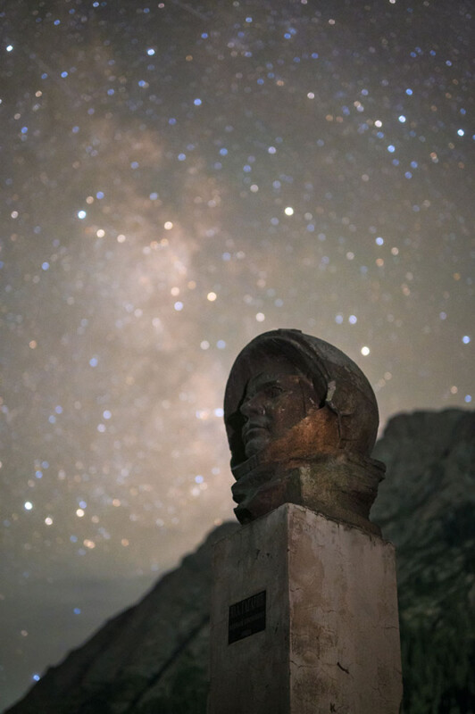 A bronze bust on a pedestal stands under a starry night sky, with galaxies and constellations clearly visible. The bust appears to be a person wearing a hood or helmet. A large mountain range is faintly visible in the background.