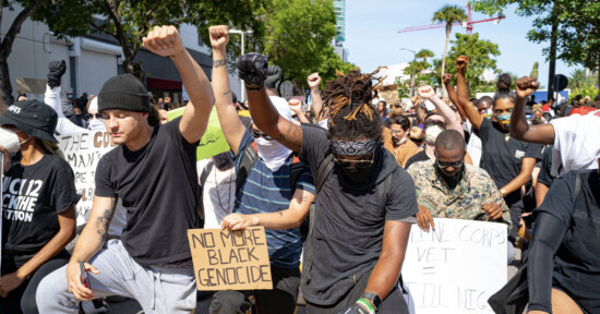 A group of protesters, diverse in age and ethnicity, kneels in solidarity with their fists raised. Some hold signs with messages such as "No More Black Genocide" and "Marine Corps Vet = Willing to Die." The atmosphere is tense and passionate.