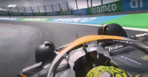 A close-up of a Formula 1 car from a first-person perspective, navigating a corner on a racetrack. The driver wears a brightly colored helmet, and sponsor logos, including "7-Eleven," are visible on the vehicle. The track is wet, and "aramco" branding is seen on barriers.