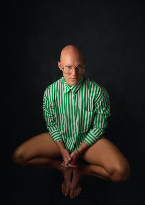 A bald person with glasses poses in front of a dark background. They are wearing a long-sleeve green and white vertically striped shirt and no visible pants, sitting cross-legged on a wooden stool, looking directly at the camera with a serious expression.