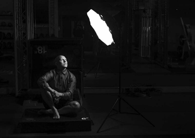 A person with a shaved head, wearing a long-sleeved button-up shirt, sits cross-legged on a raised platform in a dimly lit room, illuminated by a large softbox light. The background features gym equipment and mirrors.