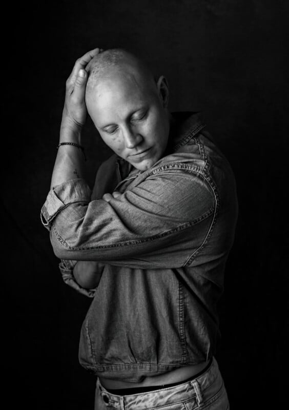 Black and white portrait of a bald person with closed eyes, wearing a denim shirt. One arm is bent, resting on top of their head, and the other arm is crossed over their chest. They appear to have a calm and reflective expression. Background is dark and plain.