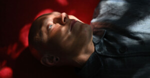 A person with a shaved head is lying down, gazing upwards. They are illuminated by a soft light, casting dramatic shadows on their face. They are wearing a striped button-up shirt. The background is dark red, adding to the moody atmosphere.