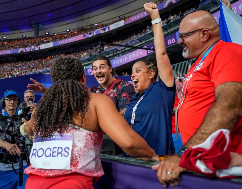 A female athlete, wearing a uniform with the name "Rogers" on the back, is celebrating with her supporters in the stands of a stadium. The supporters look excited and are cheering energetically, with one person raising their fist in the air. The stadium is filled with spectators.