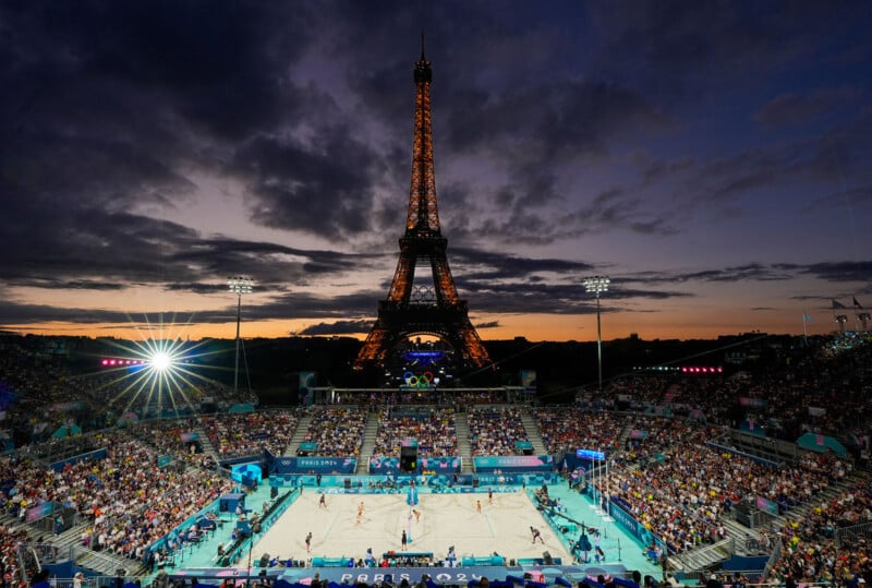 A beach volleyball match is taking place at a packed, brightly lit stadium with the Eiffel Tower prominently illuminated in the background against a partially cloudy, sunset sky. The scene is lively with spectators watching the game.