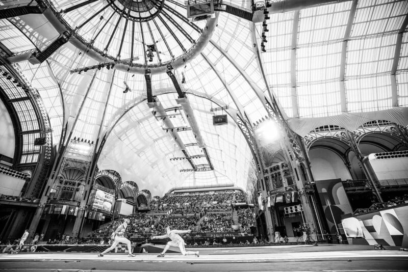 Black-and-white photo of two fencers competing in a large, ornate indoor venue with a high arched glass ceiling. The audience fills the seating area, while the foreground captures the intense duel between the fencers. The architecture is grand and symmetrical.