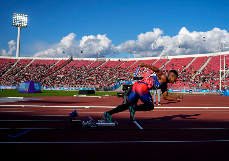 A sprinter wearing a colorful uniform bursts out of the starting blocks on a track during a race. The stadium is filled with spectators, and a scoreboard and lights are visible against a backdrop of blue sky and clouds.