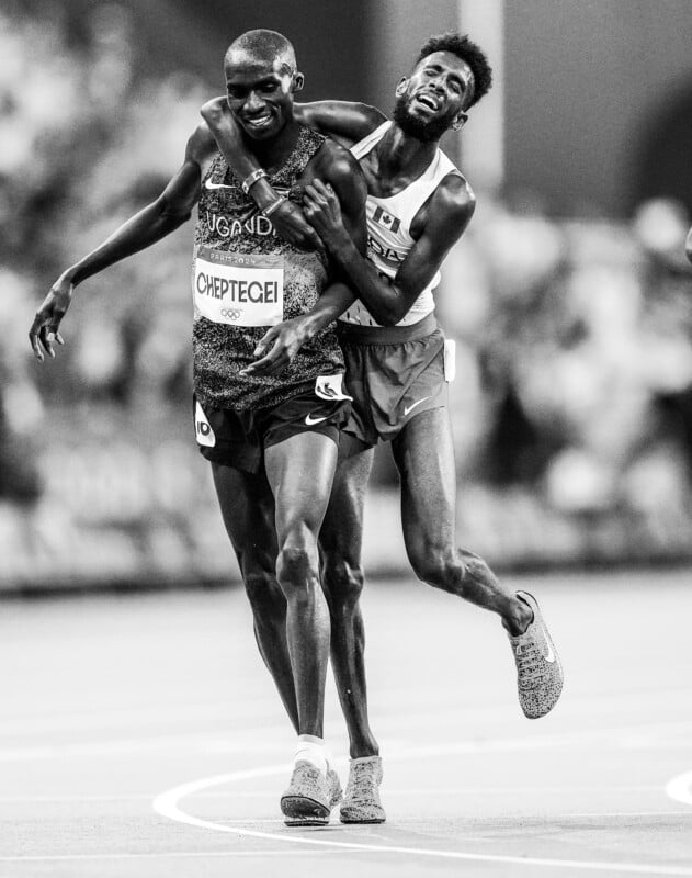 A black and white image of two athletes on a track. The athlete on the left wears a bib with the name "Cheptegei" and "Uganda". The other runner, partly obscured, has his arm around Cheptegei's shoulder as they both move forward, smiling.