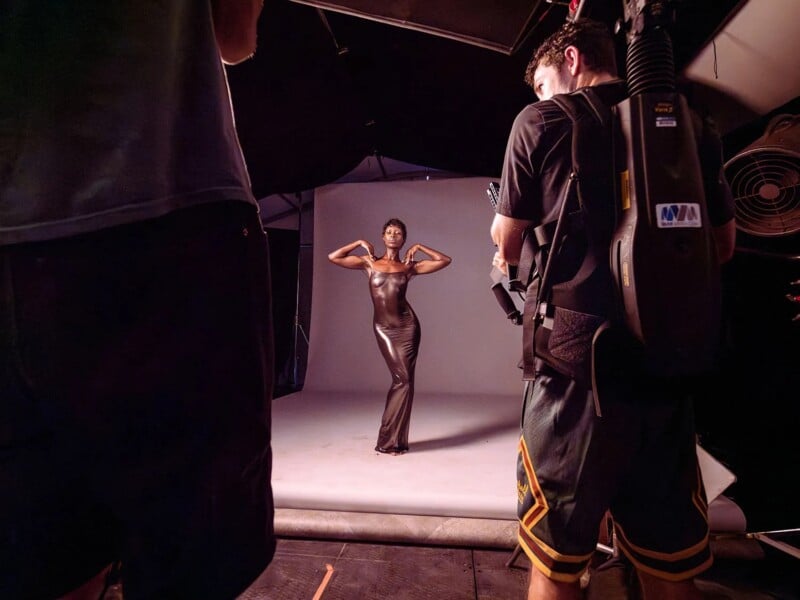 A model in a form-fitting, floor-length dress poses with her arms behind her head on a photo shoot set. She stands on a light grey backdrop, surrounded by photographers and lighting equipment. The scene is lit with studio lights.