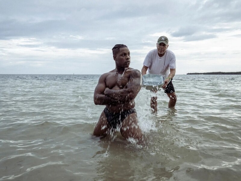 A muscular man kneels in shallow ocean water with his arms crossed, as another man wearing a baseball cap and a T-shirt behind him pours water over him from a metal container. The sky is overcast, creating a moody atmosphere.