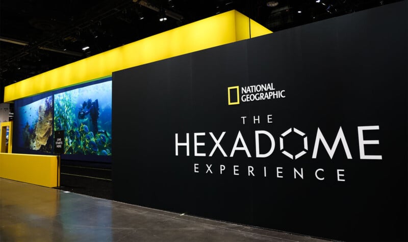 A National Geographic exhibit titled "The Hexadome Experience" is displayed at an indoor venue. The exhibit features a large sign and an illuminated section showcasing vivid underwater scenes on screens along a long wall.